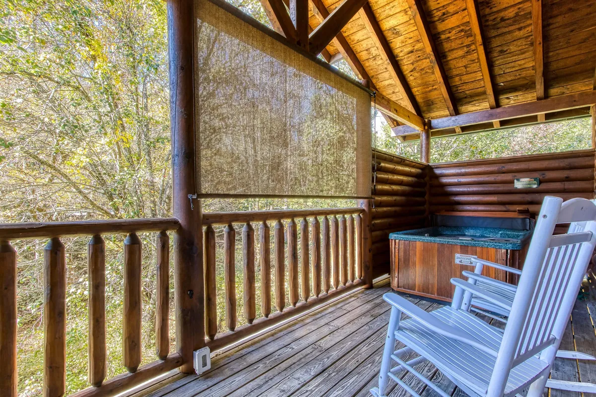 This exterior shot highlights a wooden deck featuring a hot tub, rocking chairs, and the natural surroundings. The view suggests a seamless flow between indoor comfort and outdoor relaxation, ideal for enjoying the serenity of the mountains day or night.