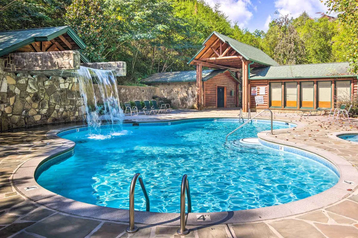 A sparkling community swimming pool with a small waterfall sits in front of a lodge-style building, offering a refreshing and scenic spot for swimming and sunbathing amidst the natural beauty of the Smokies.
