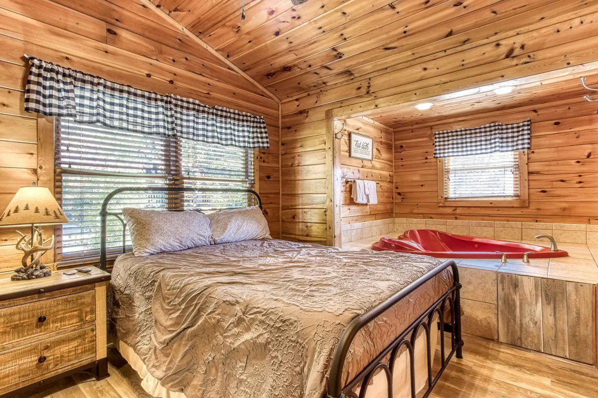 A warmly lit bedroom featuring a plush bed dressed in soft linens sits beside a deep-soaking jacuzzi tub. Wooden walls and a decorative lamp create a cozy, inviting atmosphere, while morning light filters in through a nearby window, highlighting the space's tranquil comfort and romantic design.