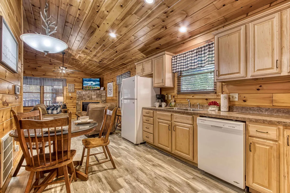 A spacious interior with a wooden ceiling, kitchen cabinetry, a dining table set against windows with checkered curtains, and a television. The dining area is positioned to encourage conversation, and the kitchen is easily accessible—ideal for socializing while cooking.