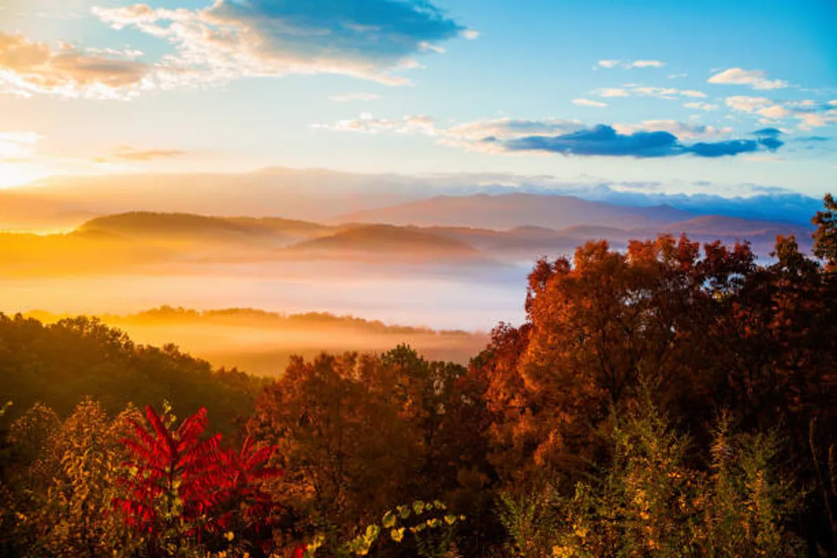 A breathtaking view of distant mountains, layered valleys filled with vibrant autumn foliage, and a colorful sunset sky. The scene conveys the tranquility and majesty of the Smokies, as seen from the property's vantage point.