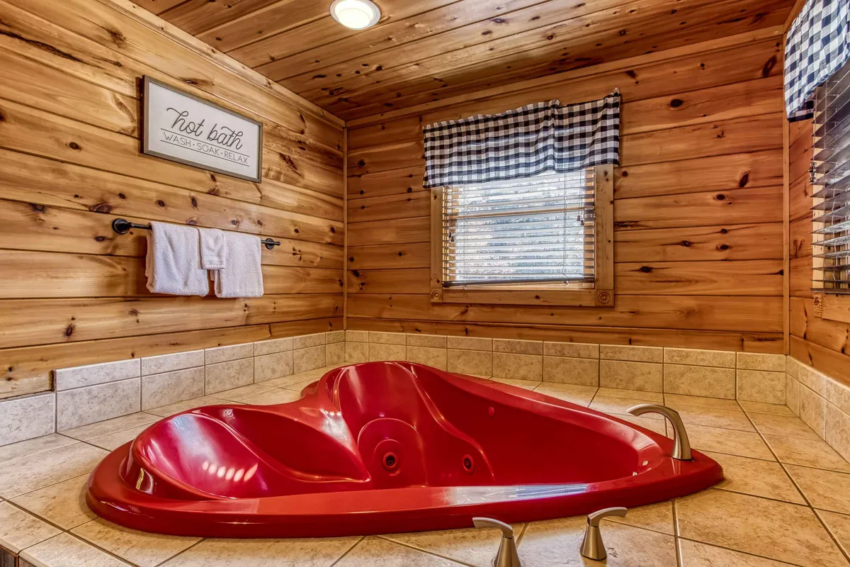A close-up of a heart-shaped jacuzzi tub set against wooden paneling, with fluffy towels on a rack and delicate, checked window treatments nearby. The warm lighting and inviting textures promise an intimate, luxurious soak ideal for romantic evenings or pampering self-care moments.