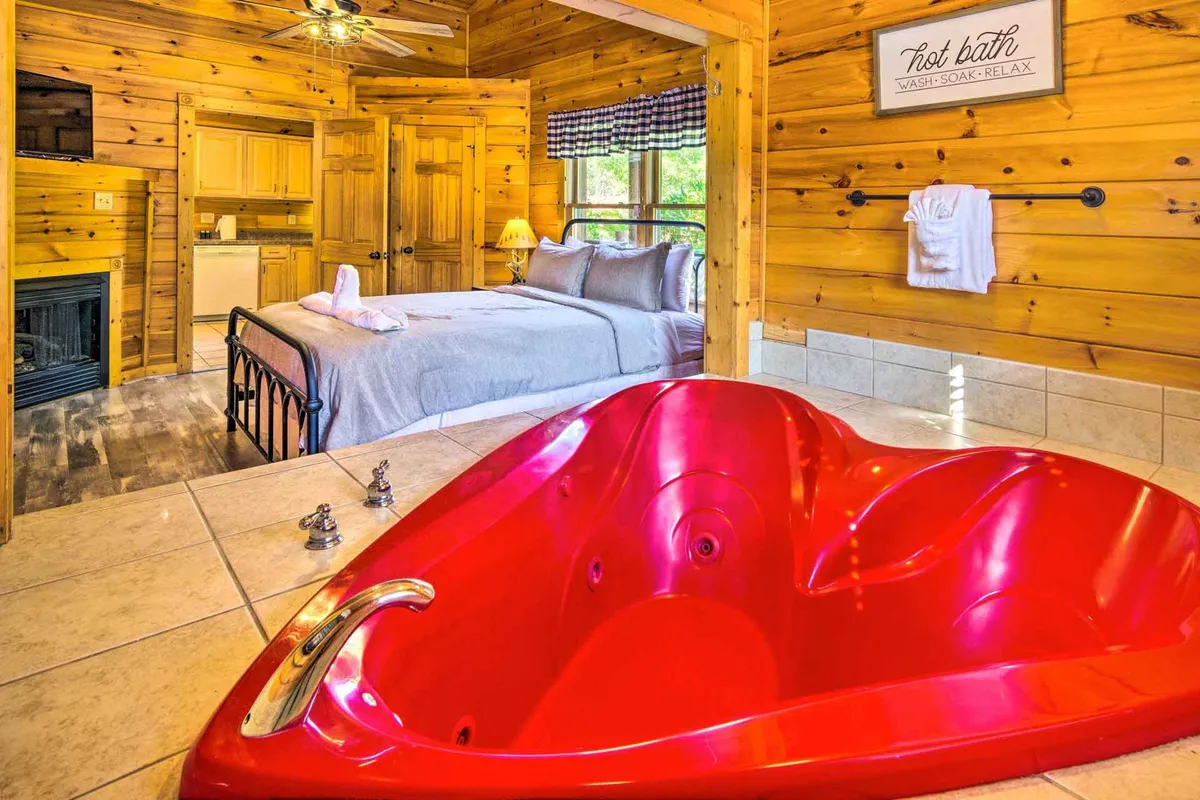This image showcases a heart-shaped hot tub set next to a king-sized bed, with warm wooden walls creating an intimate, cabin-like feel. A fireplace glows nearby, and a kitchenette is tucked at the side, reinforcing the sense of a self-contained, romantic retreat. The soft lighting and cozy decor emphasize privacy and togetherness, perfect for couples seeking a special escape.