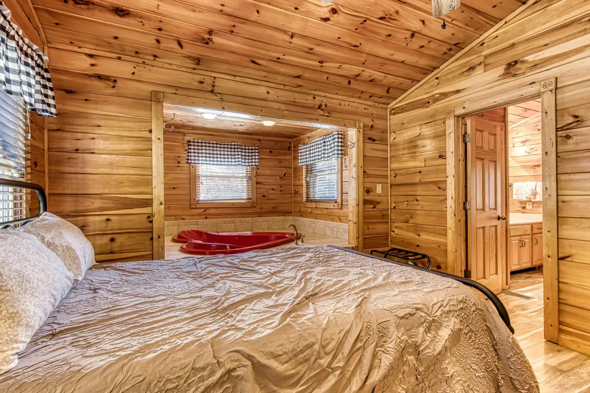A cozy cabin interior featuring wooden walls, a plush bed adorned with decorative pillows, a luxurious jacuzzi tub, and soft window treatments. The ambiance is serene and private, designed for ultimate comfort and relaxation.